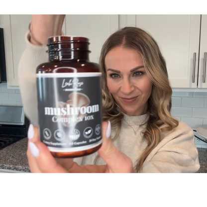 Woman holding Leah Rose Essentials Mushroom Complex 10X supplement bottle in a modern kitchen, showcasing a natural mushroom supplement for focus, energy, immune support, and daily wellness.